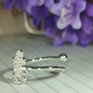 K&ouml;nnte beinhalten: Ein silberfarbener Ring mit floralem Design, besetzt mit klaren Kristallen. Der offene Ring hat eine kleine Kugel am gegen&uuml;berliegenden Ende des floralen Designs. Im Hintergrund sind unscharfe lila Blumen zu sehen.