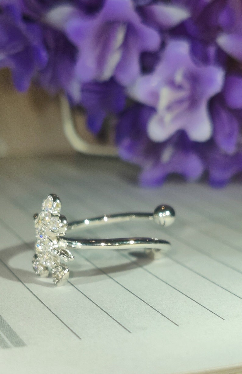 K&ouml;nnte beinhalten: Ein silberfarbener Ring mit floralem Design, besetzt mit klaren Kristallen. Der offene Ring hat eine kleine Kugel am gegen&uuml;berliegenden Ende des floralen Designs. Im Hintergrund sind unscharfe lila Blumen zu sehen.