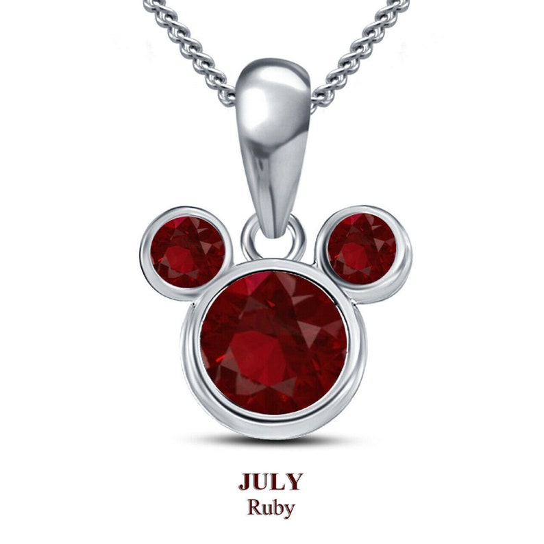May include: A silver pendant necklace featuring a Mickey Mouse-inspired design with three round, bezel-set, deep red ruby gemstones. The pendant hangs from a delicate silver chain. The text "JULY Ruby" is displayed below the pendant.