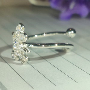K&ouml;nnte beinhalten: Ein silberfarbener Ring mit floralem Design, besetzt mit kleinen, klaren Edelsteinen. Der Ring hat ein offenes Design mit einem kleinen Ball am Ende des Bandes. Der Ring liegt auf einem linierten Papier.