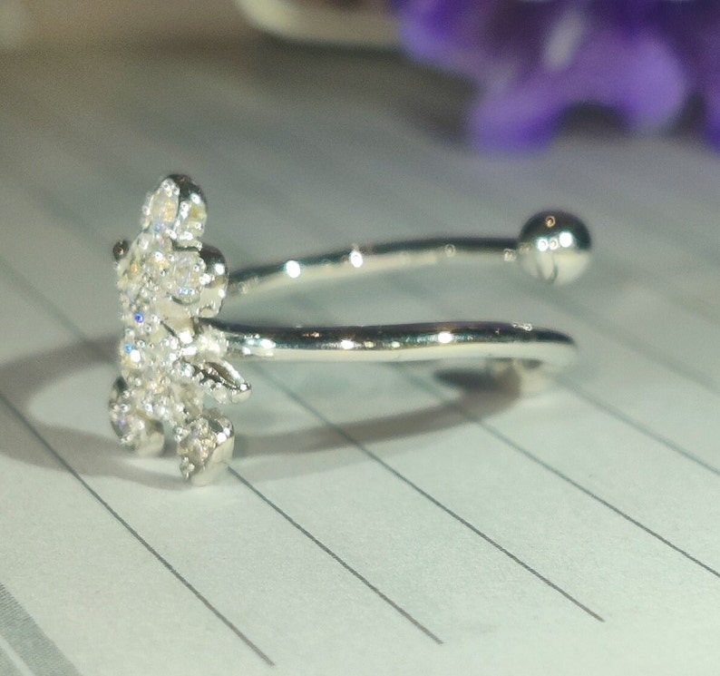 K&ouml;nnte beinhalten: Ein silberfarbener Ring mit floralem Design, besetzt mit kleinen, klaren Edelsteinen. Der Ring hat ein offenes Design mit einem kleinen Ball am Ende des Bandes. Der Ring liegt auf einem linierten Papier.