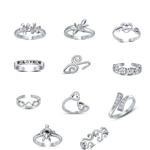 Puede incluir: Colección de anillos de dedo plateados con varios diseños. Los estilos incluyen patrones florales, de corazón y abstractos, así como un anillo con la palabra "LOVE". Algunos anillos presentan piedras preciosas transparentes. Los anillos se muestran sobre un fondo blanco.