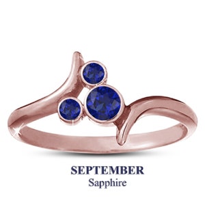 May include: Rose gold ring with three round, deep blue sapphires. The ring has a unique, flowing design with the text "SEPTEMBER Sapphire" below the ring.
