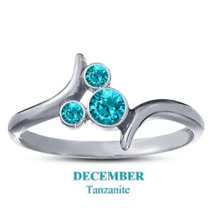 May include: A silver ring with three round, teal-colored Tanzanite gemstones. The ring has a unique, flowing design with the text "DECEMBER Tanzanite" at the bottom. The gemstones are set in a cluster arrangement.