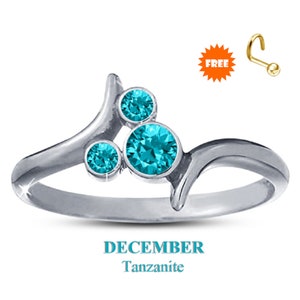 May include: A silver ring with three round, teal-colored Tanzanite stones. The ring has a unique, flowing design. The word "DECEMBER" and "Tanzanite" are displayed below the ring. A small, gold-colored nose ring is also shown.