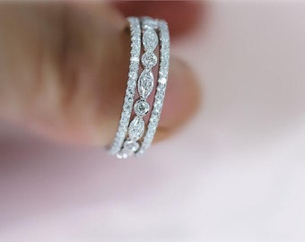 Puede incluir: Primer plano de tres anillos plateados con brillantes gemas transparentes. Los anillos presentan un diseño delicado con pequeñas piedras redondas y piedras más grandes en forma de marquesa. Fondo rosa suave.