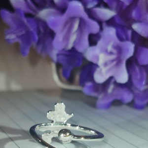 K&ouml;nnte beinhalten: Verstellbarer Ring in Silber mit kleinem Kronen-Design. Der Ring hat zwei kleine, runde Kugelakzente an den offenen Enden. Der Hintergrund zeigt verschwommene lila Blumen.