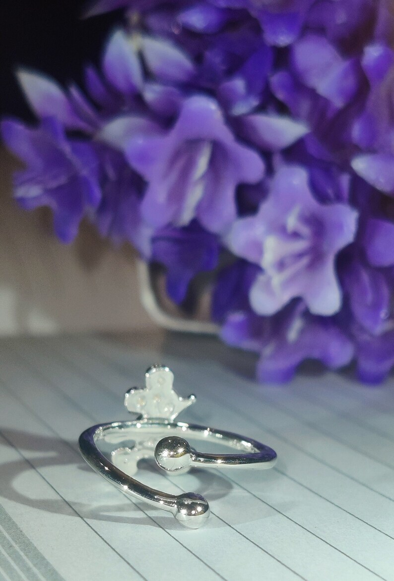 K&ouml;nnte beinhalten: Verstellbarer Ring in Silber mit kleinem Kronen-Design. Der Ring hat zwei kleine, runde Kugelakzente an den offenen Enden. Der Hintergrund zeigt verschwommene lila Blumen.