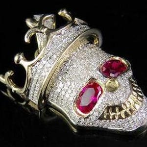 Puede incluir: Un colgante de calavera dorada con corona, incrustado con pequeñas piedras blancas brillantes. Dos gemas rojas ovaladas forman los ojos. El colgante presenta un diseño detallado sobre un fondo oscuro.