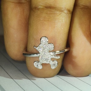 K&ouml;nnte beinhalten: Ein silberner Ring mit einem Mickey-Mouse-Motiv. Die Mickey-Mouse-Figur ist mit kleinen, funkelnden Steinen besetzt. Der Ring wird an einem Finger gezeigt, um die Details des Designs hervorzuheben.