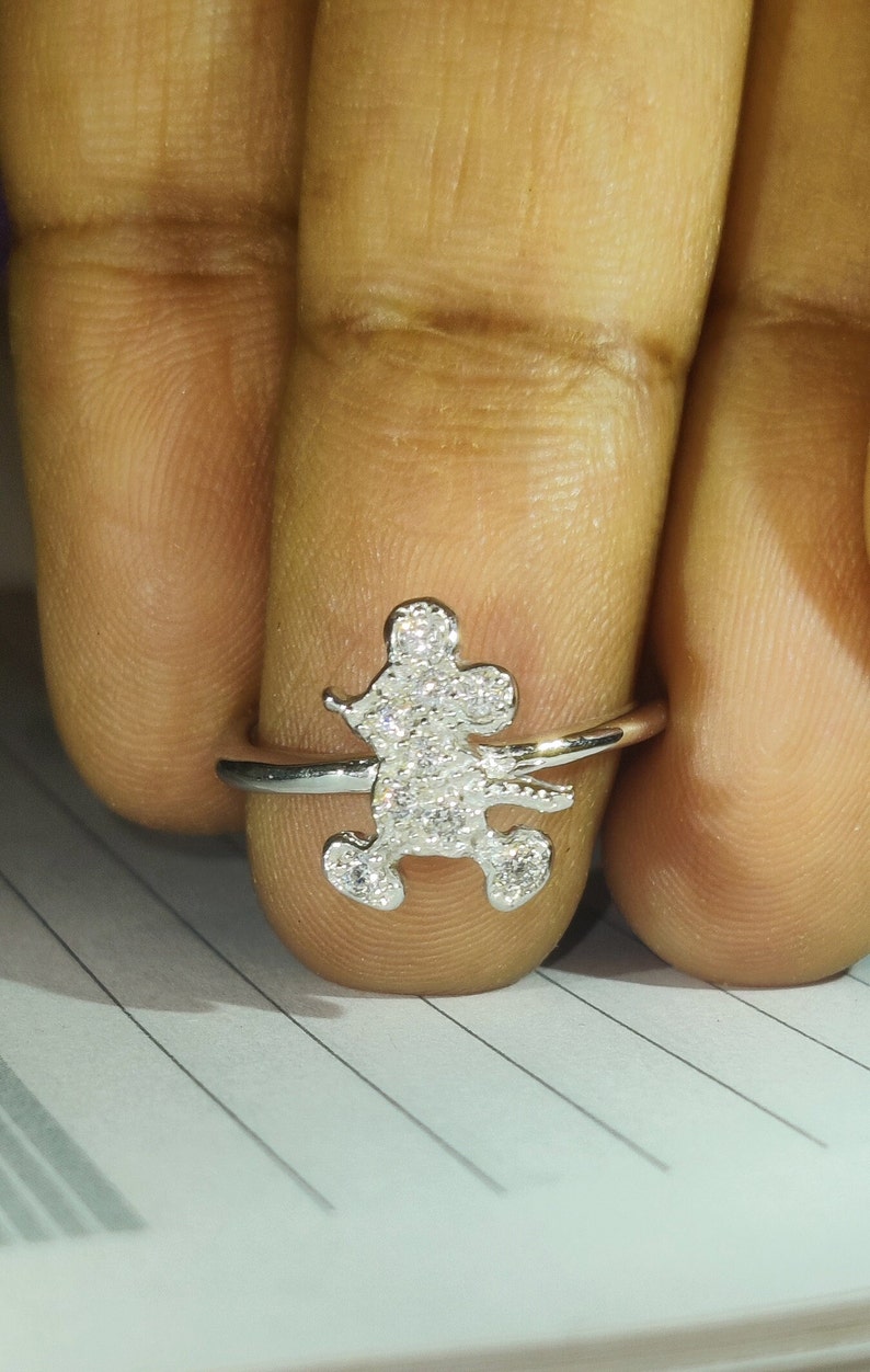 K&ouml;nnte beinhalten: Ein silberner Ring mit einem Mickey-Mouse-Motiv. Die Mickey-Mouse-Figur ist mit kleinen, funkelnden Steinen besetzt. Der Ring wird an einem Finger gezeigt, um die Details des Designs hervorzuheben.