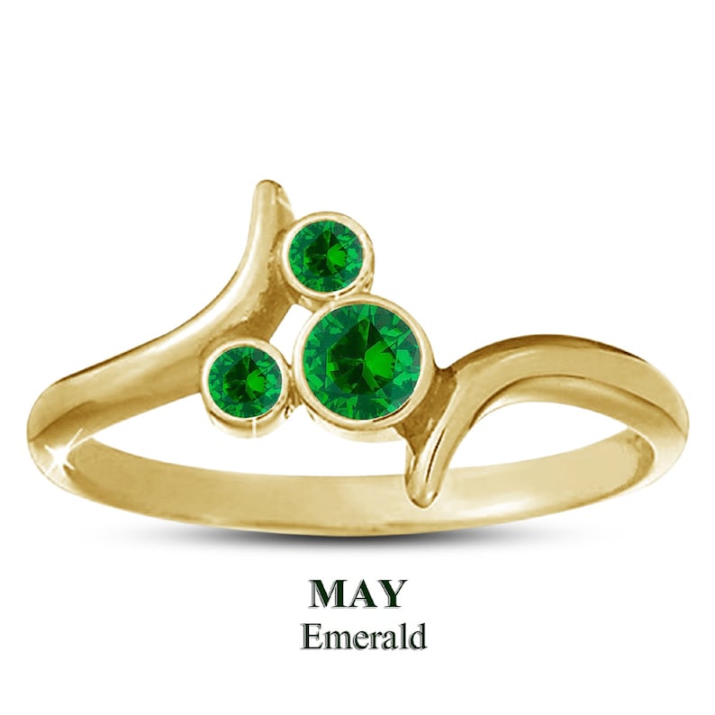 Puede incluir: Anillo dorado con tres esmeraldas verdes redondas. El dise&ntilde;o del anillo es fluido y original. El texto "MAY Emerald" es visible en la parte inferior. Las piedras est&aacute;n engastadas en un engaste de bisel.