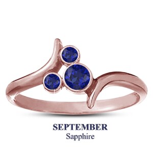 May include: Rose gold ring with three round, deep blue sapphires. The ring has a unique, flowing design with the text "SEPTEMBER Sapphire" below the ring.