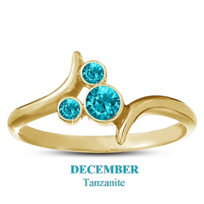 May include: A gold-colored ring with three round, bright blue gemstones. The ring has a unique, flowing design with the text "DECEMBER Tanzanite" below the ring.