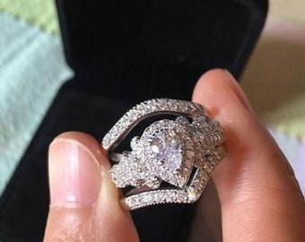 Puede incluir: Un juego de anillos de plata de tres piezas con una piedra central grande en forma de pera rodeada de piedras más pequeñas. El conjunto incluye una banda a juego y una banda curva, todas brillantes con piedras preciosas transparentes. Los anillos se muestran en una caja negra.