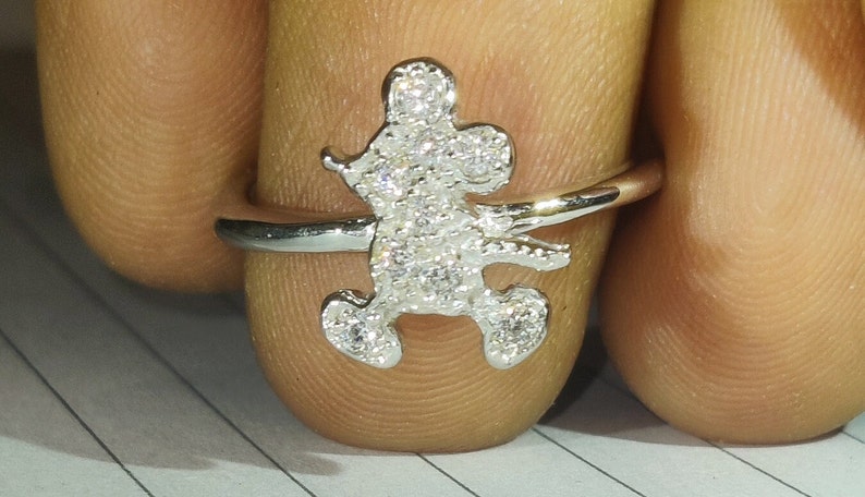 K&ouml;nnte beinhalten: Ein silberner Ring mit einem Mickey-Mouse-Motiv. Die Mickey-Mouse-Figur ist mit kleinen, funkelnden Kristallen besetzt. Der Ring wird an einem Finger gezeigt, um die Details des Designs hervorzuheben.