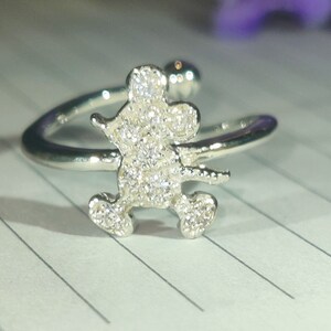 K&ouml;nnte beinhalten: Silberfarbener Ring mit einer glitzernden, mit Edelsteinen besetzten Figur einer Zeichentrickmaus. Der Ring hat ein offenes Design, mit der Mausfigur in der Mitte und einem gebogenen Band, das sich um den Finger legt. Der Ring ist vor einem linierten Papierhintergrund platziert.