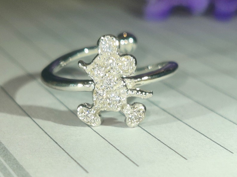 K&ouml;nnte beinhalten: Silberfarbener Ring mit einer glitzernden, mit Edelsteinen besetzten Figur einer Zeichentrickmaus. Der Ring hat ein offenes Design, mit der Mausfigur in der Mitte und einem gebogenen Band, das sich um den Finger legt. Der Ring ist vor einem linierten Papierhintergrund platziert.