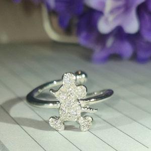 K&ouml;nnte beinhalten: Silberfarbener Ring mit einem Mickey-Mouse-Design, besetzt mit funkelnden, klaren Kristallen. Der Ring hat ein offenes, verstellbares Design. Der Hintergrund zeigt verschwommene lila Blumen und eine karierte Papieroberfl&auml;che.
