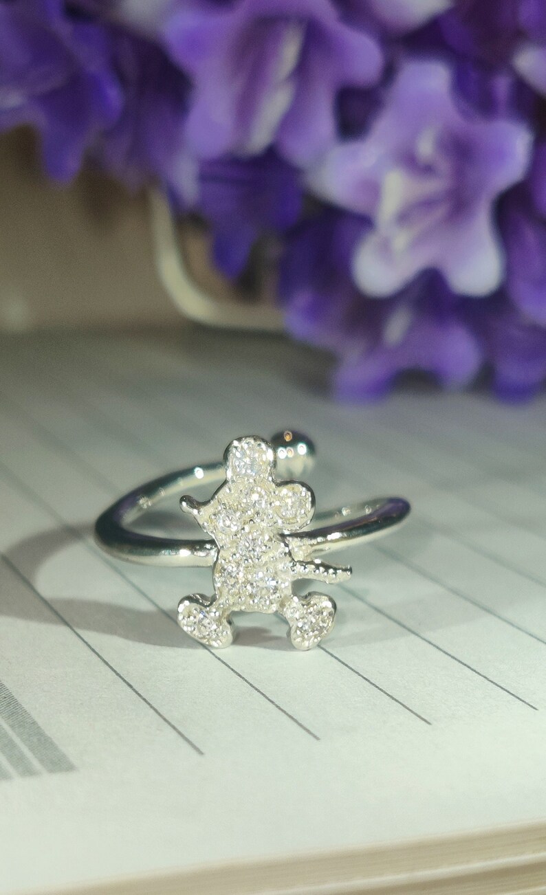 K&ouml;nnte beinhalten: Silberfarbener Ring mit einem Mickey-Mouse-Design, besetzt mit funkelnden, klaren Kristallen. Der Ring hat ein offenes, verstellbares Design. Der Hintergrund zeigt verschwommene lila Blumen und eine karierte Papieroberfl&auml;che.
