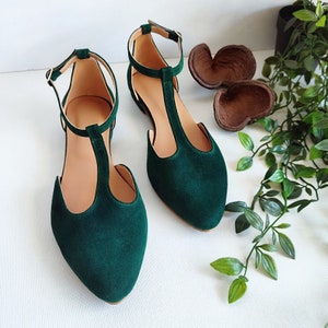 May include: A pair of emerald green T-strap flats with pointed toes. The shoes have a delicate ankle strap with a small gold buckle. The interior of the shoe is a light tan color. The shoes are displayed on a white surface.