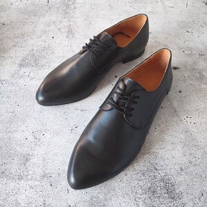 May include: A pair of black leather dress shoes with a pointed toe and lace-up closure. The shoes have a smooth, polished finish and a light brown interior. The shoes are displayed on a gray, textured surface.