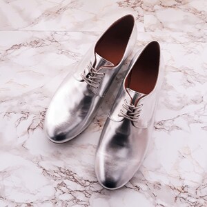 May include: A pair of silver leather lace-up shoes on a white marble background.