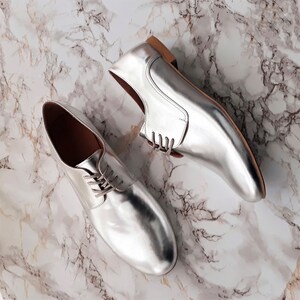May include: A pair of silver leather lace-up shoes with a pointed toe. The shoes have a light brown sole and are photographed on a white marble surface.