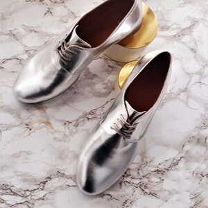 May include: A pair of silver metallic leather oxfords with lace-up closures. The shoes are on a white marble surface.