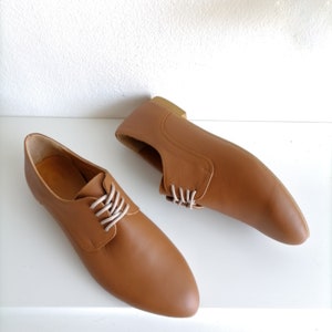 May include: A pair of brown leather shoes with white laces. The shoes have a classic design with a rounded toe and a low heel. The shoes are displayed on a white surface.