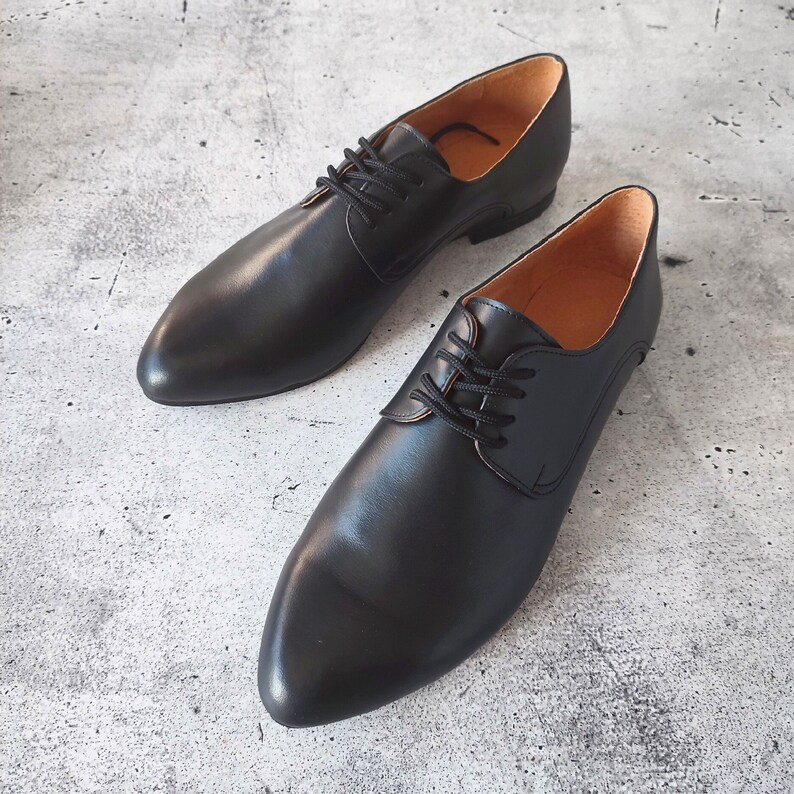 May include: A pair of black leather dress shoes with a pointed toe and lace-up closure. The shoes have a smooth, polished finish and a light brown interior. The shoes are displayed on a gray, textured surface.
