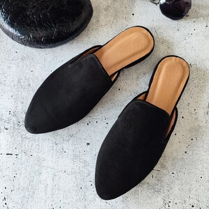 K&ouml;nnte beinhalten: Zwei schwarze Wildlederslipper mit spitzer Zehenpartie. Die Slipper haben eine braune Ledersohle.