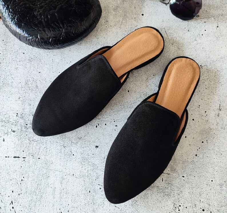 K&ouml;nnte beinhalten: Zwei schwarze Wildlederslipper mit spitzer Zehenpartie. Die Slipper haben eine braune Ledersohle.