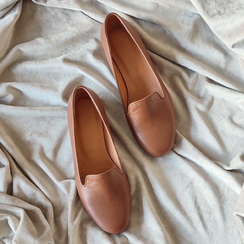 Puede incluir: Un par de mocasines de cuero marr&oacute;n. Los zapatos son de color marr&oacute;n claro y tienen un dise&ntilde;o cl&aacute;sico sin cordones. Los mocasines se exhiben sobre una tela gris claro texturizada.
