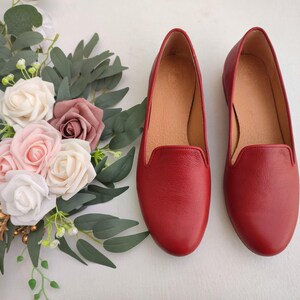 Puede incluir: Un par de zapatos sin cordones de cuero rojo con punta redondeada. Los zapatos est&aacute;n junto a un arreglo floral de rosas blancas, rosas y malvas con hojas verdes. Los zapatos tienen un interior beige.