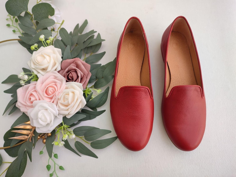 Puede incluir: Un par de zapatos sin cordones de cuero rojo con punta redondeada. Los zapatos est&aacute;n junto a un arreglo floral de rosas blancas, rosas y malvas con hojas verdes. Los zapatos tienen un interior beige.