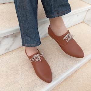 May include: A pair of brown leather oxford shoes with light-colored laces. The shoes have a pointed toe and are shown from a top-down perspective. The shoes are on a set of stairs, and the person wearing them is wearing blue jeans.