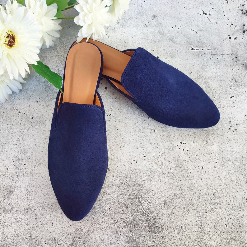May include: A pair of navy blue suede slip-on mules with pointed toes and tan insoles. The shoes are displayed on a gray surface with white flowers in the background. These backless shoes are a stylish footwear option.
