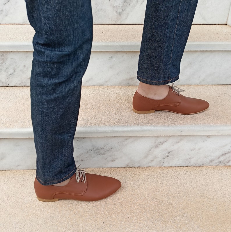 May include: A pair of brown leather dress shoes with light-colored laces. The shoes are worn with blue denim jeans. The image shows a person walking up a set of stairs.
