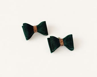 May include: Two dark green velvet hair bows, each featuring a central rectangular metallic accent piece with small embedded crystals. The bows have a soft, textured fabric finish and are positioned diagonally against a plain white background.
