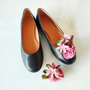 May include: A pair of black leather ballet flats with pink flower embellishments. The flowers are arranged on the top of the shoes and are made of fabric.