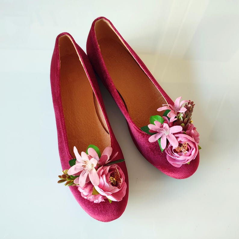 May include: A pair of burgundy velvet flats with pink flower embellishments. The flowers are arranged in a cluster on the toe of each shoe.