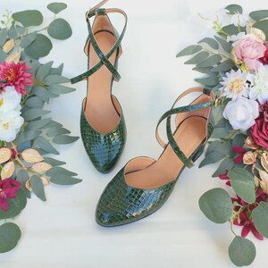 May include: A pair of green, textured, pointed-toe heels with ankle straps. The shoes have a criss-cross design over the foot and a small heel. The background features floral arrangements with pink, white, and burgundy flowers, and green foliage.