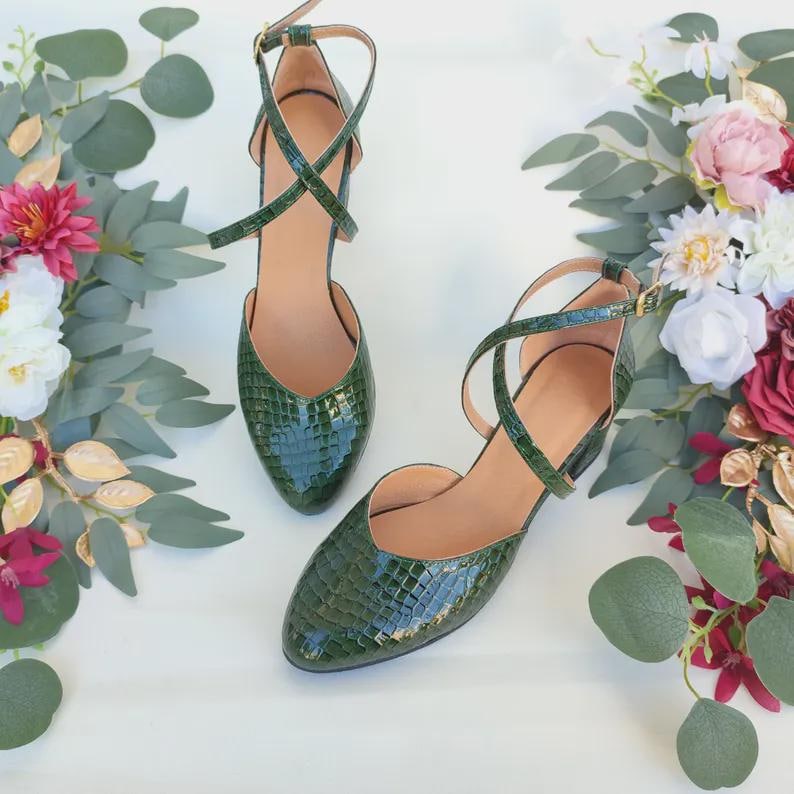 May include: A pair of green, textured, pointed-toe heels with ankle straps. The shoes have a criss-cross design over the foot and a small heel. The background features floral arrangements with pink, white, and burgundy flowers, and green foliage.