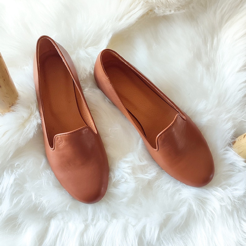 Puede incluir: Un par de mocasines de cuero marr&oacute;n. Los zapatos tienen una punta redondeada y un perfil bajo. El cuero tiene un acabado liso y un color marr&oacute;n rojizo c&aacute;lido. Los zapatos se exhiben sobre una superficie blanca y esponjosa.