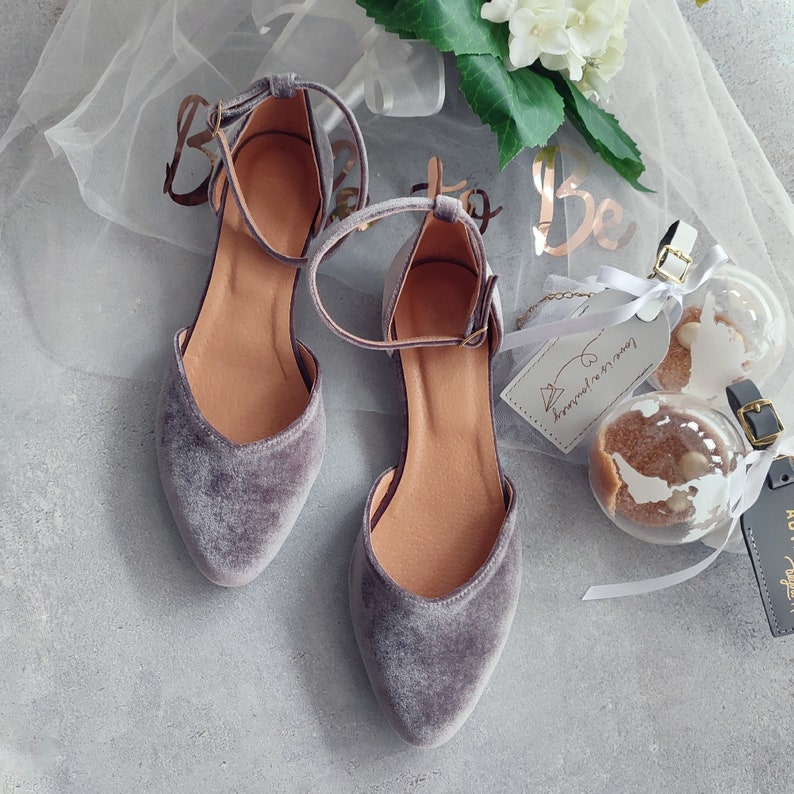 May include: A pair of grey velvet ankle strap shoes with a pointed toe. The shoes are on a white background with a tulle overlay and a decorative tag that says "Love is in the air".