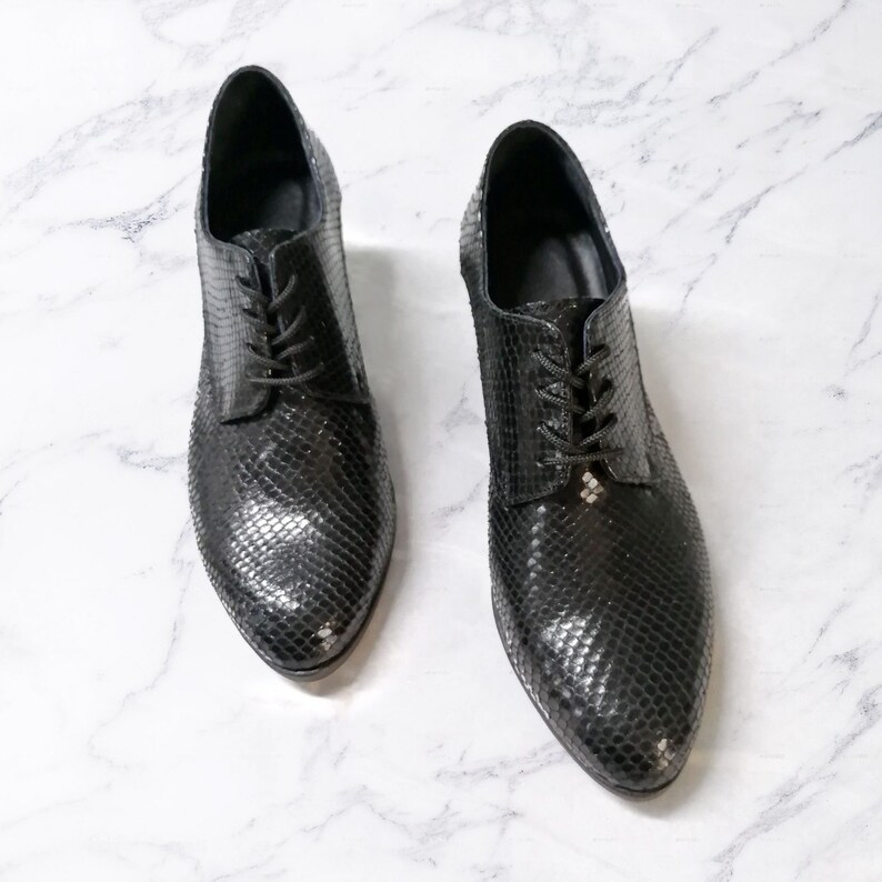 May include: A pair of black, textured leather oxford shoes with laces. The shoes have a pointed toe and a low heel. The leather has a scale-like pattern. The shoes are displayed on a white marble surface.