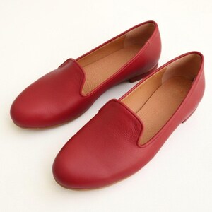 Puede incluir: Un par de mocasines de cuero rojo. Los zapatos tienen un dise&ntilde;o cl&aacute;sico sin cordones con punta redondeada y tac&oacute;n bajo. El interior es de color marr&oacute;n claro y los zapatos est&aacute;n sobre un fondo blanco.