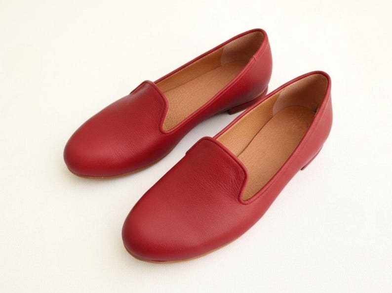 Puede incluir: Un par de mocasines de cuero rojo. Los zapatos tienen un dise&ntilde;o cl&aacute;sico sin cordones con punta redondeada y tac&oacute;n bajo. El interior es de color marr&oacute;n claro y los zapatos est&aacute;n sobre un fondo blanco.
