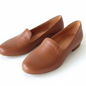 Puede incluir: Un par de mocasines de cuero marr&oacute;n. Los zapatos tienen una punta redondeada y un tac&oacute;n bajo. El cuero parece liso y los zapatos son de color marr&oacute;n claro. Los zapatos se muestran sobre un fondo blanco.